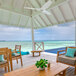A white Haiku outdoor ceiling fan on a white ceiling above a table and chairs on a wooden deck overlooking the ocean.
