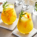 Two glasses of orange juice with rosemary sprigs on a table with white coasters.