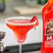 A glass of Jose Cuervo Strawberry Margarita with a strawberry on the rim next to a red bottle of Jose Cuervo Strawberry Margarita.