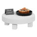 A Cal-Mil white wood round display stand holding a plate of food on a small white table.