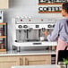 An Astra Mega II M2S-017-WH semi-automatic white espresso machine on a countertop with a person preparing a coffee.