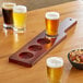 An Acopa mahogany flight paddle holding glasses of beer and a bowl of mixed nuts on a table.