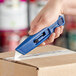 A hand holding a blue Allway Tools plastic retractable utility knife to cut a box.