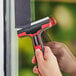A person using an Allway Tools glass and tile scraper with a red and black handle to clean a window.