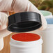 A hand in gloves holding a plastic container with a black 38/400 smooth lid.
