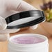 A hand holding a 70/400 smooth black lid over a white container with purple liquid.