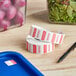 A roll of white and red Noble Products Wednesday date labels on a blue tray next to a container of salad.