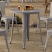 A Lancaster Table & Seating Alloy Series round nickel gray outdoor table with chairs and glasses of beer on a stone surface.