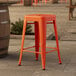 A Lancaster Table & Seating outdoor backless counter height stool in amber orange on a stone patio.