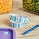 A roll of white Noble Products Monday labels next to a blue pen and a purple container of salad.