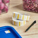 A blue tray with a roll of white and yellow Noble Products Tuesday 1" Dissolvable Day of the Week Labels and a pen next to a container of food.