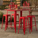 A Lancaster Table & Seating ruby red metal table with four backless barstools on an outdoor patio.