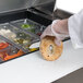 A person in gloves using a knife to cut a bagel on a stainless steel counter over a Traulsen sandwich prep table.