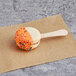 A Coco Bakery salted caramel macaron pop on a wooden stick with orange icing and sprinkles.