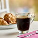 A glass cup of Green Mountain Coffee Roasters Dark Magic coffee on a table with croissants.