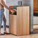 A woman putting a bottle of water into a Lancaster Table & Seating weathered walnut trash can with a "THANK YOU" swing door.