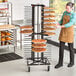 A man in an apron standing next to a JackStack mobile pizza plate rack filled with pizzas.