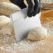 A person in black gloves uses a Choice stainless steel dough cutter with a white handle to cut white dough on a brown table.