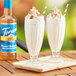 Two glasses of milkshakes with straws and a bottle of Torani Sugar-Free Peanut Butter Cup flavoring syrup on a wood table.