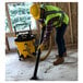 A man in a yellow safety vest and hard hat using a yellow and black DeWalt wet/dry vacuum to clean a wood floor.