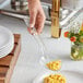 A hand holding a Choice clear polycarbonate salad bar spoon over a plate of macaroni and cheese.