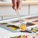 A person holding a Choice clear polycarbonate salad bar spoon over a bowl of food.