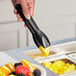 A hand using black polycarbonate salad tongs to serve fruit into a bowl.