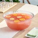 A clear plastic punch bowl of pink liquid with lemons and slices of oranges on a table.