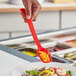 A hand holding a red Choice polycarbonate salad spoon over a salad.