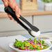 A person using Choice black flat grip tongs to serve a salad
