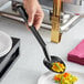 A hand holding a black Choice Perforated Salad Bar Spoon scooping food onto a plate.