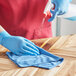 A person wearing blue gloves cleaning a wooden table with a 3M Scotch-Brite blue microfiber cloth.