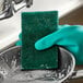 A gloved hand using a 3M Scotch-Brite heavy-duty green sponge to clean a pan.