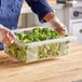 A person wearing gloves holding a Choice clear polycarbonate food storage box with broccoli inside.