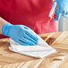 A person wearing blue gloves cleaning a wood table with a 3M Scotch-Brite white microfiber cloth.