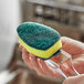 A hand using a green and yellow 3M Scotch-Brite Heavy-Duty scrub pad to clean a glass.