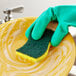 A gloved hand using a 3M Scotch-Brite heavy-duty scrub sponge to clean a yellow plate.