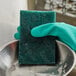 A gloved hand using a 3M Scotch-Brite heavy-duty scouring pad in a bowl.