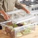 A person in a chef's jacket opening a clear plastic Choice food storage container lid to put green and red lettuce inside.