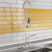 A wall-mount pre-rinse faucet with an add-on faucet, wall bracket, riser, swing nozzle, and hose installed above a stainless steel sink.