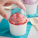 A hand holding a white paper cup with a dome lid filled with pink frozen yogurt.