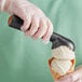 A person in a glove holding a black-handled gold ice cream scoop filled with ice cream.