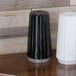 A black container of white pepper on a table with a white column and a black and white pepper shaker.