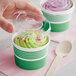 A hand holding a wooden spoon over a green paper cup of frozen yogurt with a plastic lid.