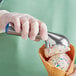 A hand in a blue glove holding a blue aluminum ice cream scoop filled with ice cream over a waffle cone.