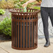 A woman standing next to a brown LT&amp;S slatted steel outdoor trash can with a flat lid.