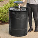 A person standing next to a Lancaster Table & Seating heavy-duty black steel outdoor trash can with a rain bonnet lid.