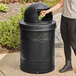 A person putting a paper cup into a Lancaster Table & Seating outdoor trash can with a plastic dome lid.