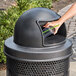 A hand putting a plastic cup with a straw into a black plastic dome lid on a trash can.