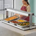 A woman in a red apron putting food into a ServIt buffet warmer on a counter.